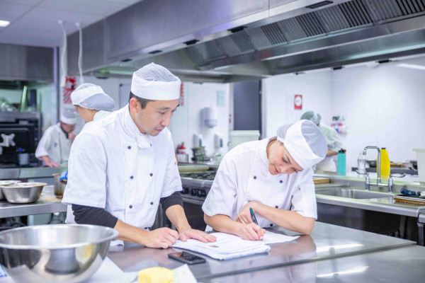 Student chefs planning with pen and paper in kitchen.